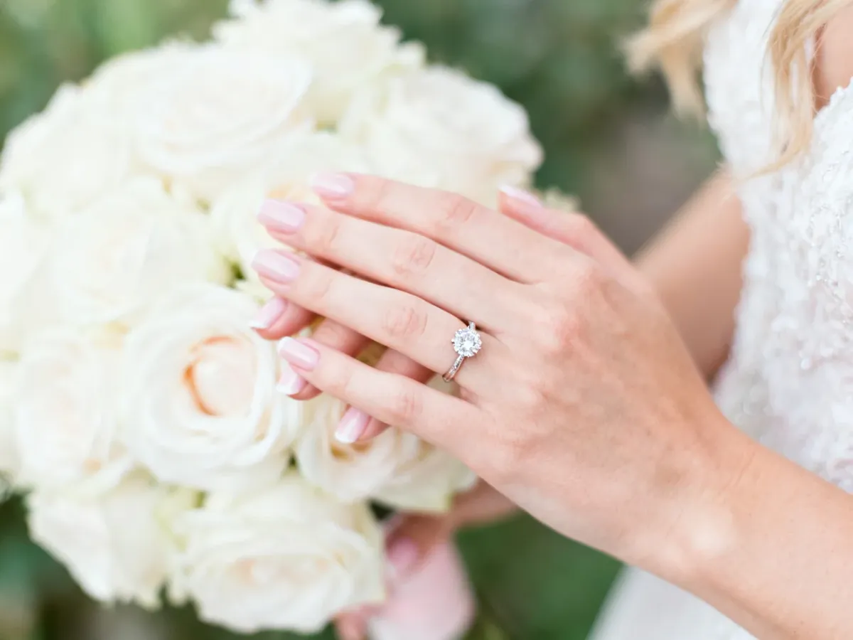 Elegancki manicure ślubny z delikatnym pierścionkiem na tle białych róż, utrzymany w romantycznej, pastelowej tonacji.